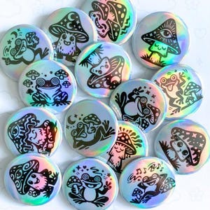May include: A collection of holographic button pins featuring black and white illustrations of frogs and mushrooms. The pins have a reflective, iridescent finish. Text on the image reads "BUTTON PINS" and "HOLO SHROOMY FROGS."