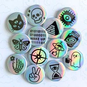 May include: A collection of holographic button pins featuring various symbols and designs. The pins include a cat, skull, eye, pyramid, jar, alien, compass, ghost, peace sign, hourglass, and a skeleton hand. The text "BUTTON PINS" and "HOLO SYMBOLS" are also visible.