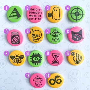 May include: A collection of fourteen neon-colored button pins with various symbols, including a pyramid, a ghost, a compass, and a cat. The text "BUTTON PINS" and "NEON SYMBOLS" are also visible. The pins are arranged on a light purple surface.