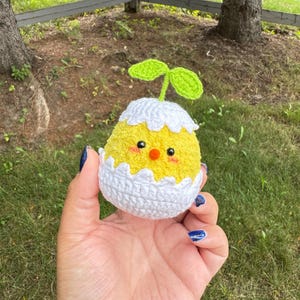 May include: A small, round, yellow crocheted chick emerging from a white crocheted eggshell. The chick has black bead eyes, a small orange beak, and pink blush marks. A green crocheted sprout with two leaves sits on top of the white eggshell.