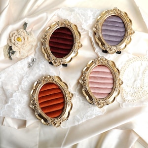 May include: Four ornate, gold-colored oval ring displays with velvet inserts in shades of red, orange, pink, and purple. The displays sit on a white satin surface with lace accents, showcasing a vintage aesthetic.