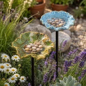 May include: Two garden stakes featuring shallow, ruffled, leaf-shaped basins in glossy blue and green ceramic. Each basin is filled with water and smooth, light-colored river stones, with a small bee resting on the green basin's edge.