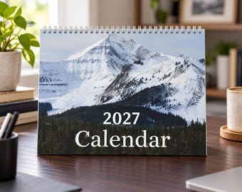 May include: A spiral-bound desk calendar for the year 2027 featuring a landscape photograph of a snow-covered mountain peak rising above a dark green pine forest. The text 2027 CALENDAR is printed in white in the center of the image.