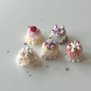 Puede incluir: Una colección de cinco mini pasteles de ganchillo en varios colores pastel. Cada pastel presenta elementos decorativos como cerezas, glaseado y lazos. Los pasteles están dispuestos sobre una superficie blanca con confeti en forma de estrella esparcido.