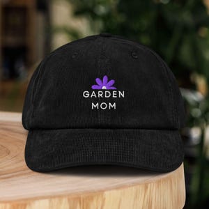 Puede incluir: Gorra de béisbol de pana negra con el texto bordado "GARDEN MOM" en blanco y un gráfico de flor morada. La gorra tiene una visera curva y una corona estructurada, perfecta para jardinería.