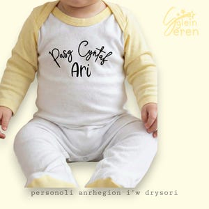 Puede incluir: Un body blanco para bebé con mangas y cuello amarillos. El body presenta el texto "Pasg Cyntaf Ari" en escritura negra. El bebé lleva el body. El texto "personoli anrhegion i'w drysori" está en la parte inferior.