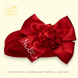 May include: A red baby headband with a large satin bow and a smaller, ruffled flower accent. The name "Nel" is written in white script on the bow. The headband is a solid red color.