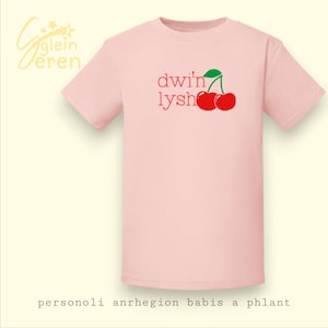 May include: A pale pink t-shirt with the words "dwi'n lysh" in red, and a graphic of two red cherries with a green leaf. The shirt has text at the bottom. This is a baby and children's apparel item.