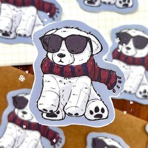 May include: A white cartoon dog sticker wearing black sunglasses and a red and blue striped scarf. The dog is sitting down with its paws visible. The sticker has a light blue border and snowflake accents.