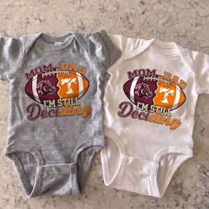 May include: Two short-sleeve baby bodysuits, one heather gray and one white, each featuring a graphic of a football split into two halves. The left side is maroon with a hog logo, the right is orange with a white T, and text reads MOM DAD I'M STILL Deciding.
