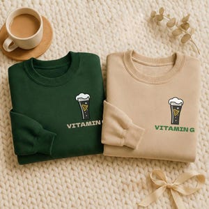 Puede incluir: Dos sudaderas de cuello redondo, una verde oscuro y otra beige, se muestran sobre una superficie texturizada de color crema. Cada sudadera presenta un gr&aacute;fico de un vaso de cerveza con el texto "VITAMIN" o "VITAMIN G" debajo. Tambi&eacute;n se ven una taza de caf&eacute; y un lazo.