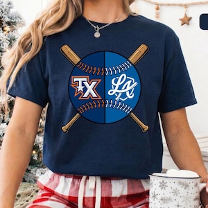 May include: Navy blue t-shirt featuring a baseball design. The design includes a baseball split in half with the letters "TX" and "LA" on each side, crossed by two baseball bats. The shirt is a comfortable fit.