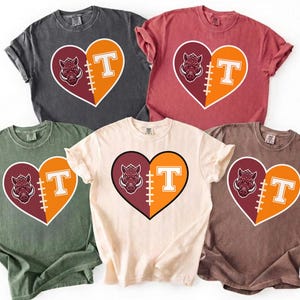 May include: Five short-sleeve t-shirts in charcoal, red, olive green, cream, and brown. Each shirt features a central heart graphic split into two halves: one side displays a maroon boar head, the other an orange letter T with football laces.