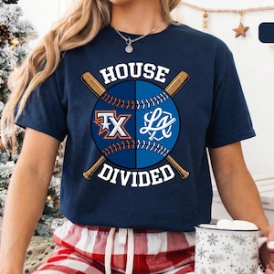 May include: Navy blue t-shirt featuring a baseball design with crossed bats. The design includes the text "HOUSE DIVIDED" and team logos "TX" and "LA" on a baseball. The shirt is a short-sleeved crew neck.