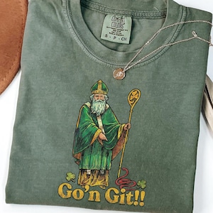 May include: Olive green t-shirt featuring a graphic of a St. Patrick's Day figure in green robes holding a staff. The text "Go'n Git!!" is below the figure. A gold necklace and a brown hat are in the background.