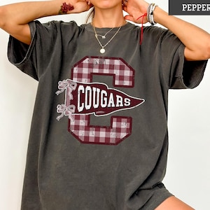 May include: A charcoal gray short-sleeve t-shirt featuring a large maroon and white gingham letter C graphic. Overlaid on the letter is a maroon pennant with the white text COUGARS and two small white bows on the left side of the design.