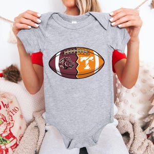 May include: A heather gray short-sleeve baby bodysuit featuring a graphic of a football split into two halves. The left side is maroon with a wild boar mascot, and the right side is orange with a white letter T. The design includes white laces.