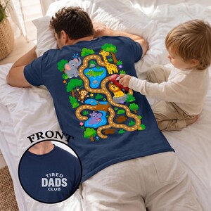 May include: Navy blue t-shirt with a colorful jungle-themed graphic on the back, featuring an elephant, giraffe, monkey, lion, and hippo along a winding road. The front of the shirt has the text "TIRED DADS CLUB" in white.
