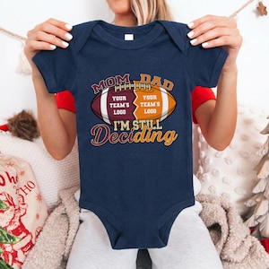 May include: Navy blue baby bodysuit with a football graphic. The graphic reads "MOM DAD" above a football design with space for team logos and the text "I'M STILL Deciding".