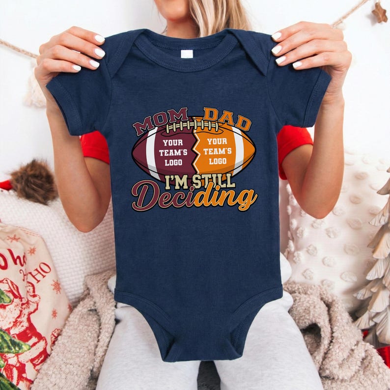 May include: Navy blue baby bodysuit with a football graphic. The graphic reads "MOM DAD" above a football design with space for team logos and the text "I'M STILL Deciding".