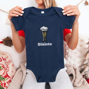 May include: Navy blue baby bodysuit with a graphic of a pint of stout beer and the word "Sl&aacute;inte" in white. The bodysuit is short-sleeved and has a round neckline. The design is centered on the front of the garment.
