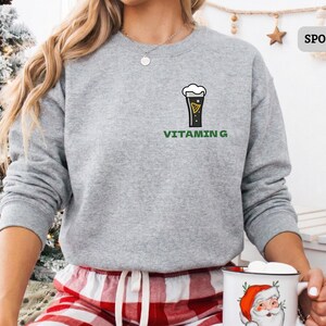 Puede incluir: Sudadera gris deportiva con un gr&aacute;fico de vaso de cerveza y el texto "Vitamin G" en verde. La sudadera de cuello redondo y manga larga se usa con un pantal&oacute;n de pijama a cuadros rojos y blancos. La imagen tambi&eacute;n muestra un &aacute;rbol de Navidad.
