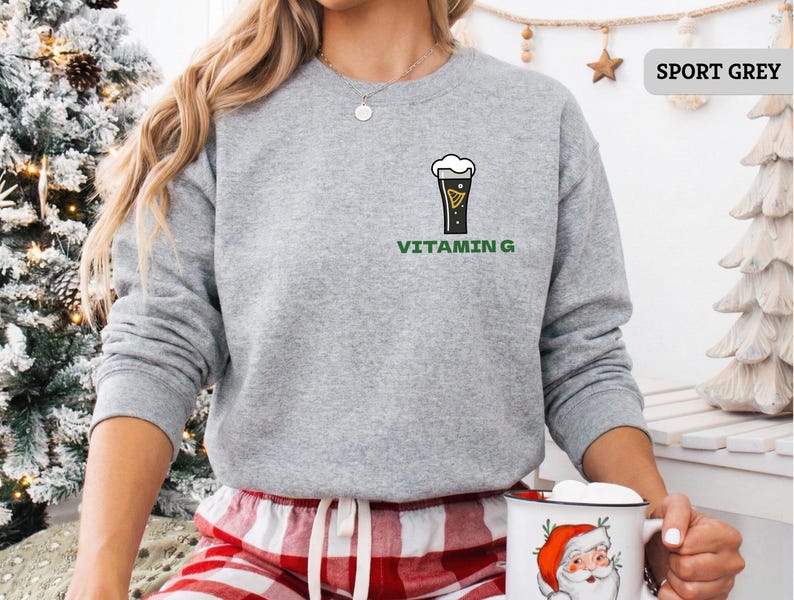 Puede incluir: Sudadera gris deportiva con un gr&aacute;fico de vaso de cerveza y el texto "Vitamin G" en verde. La sudadera de cuello redondo y manga larga se usa con un pantal&oacute;n de pijama a cuadros rojos y blancos. La imagen tambi&eacute;n muestra un &aacute;rbol de Navidad.