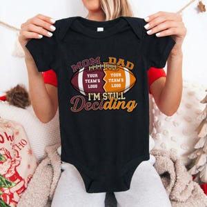 May include: Black baby bodysuit with a football graphic. The graphic reads "MOM DAD" above a football design with space for team logos and the text "I'M STILL Deciding". The bodysuit is held up by a person.