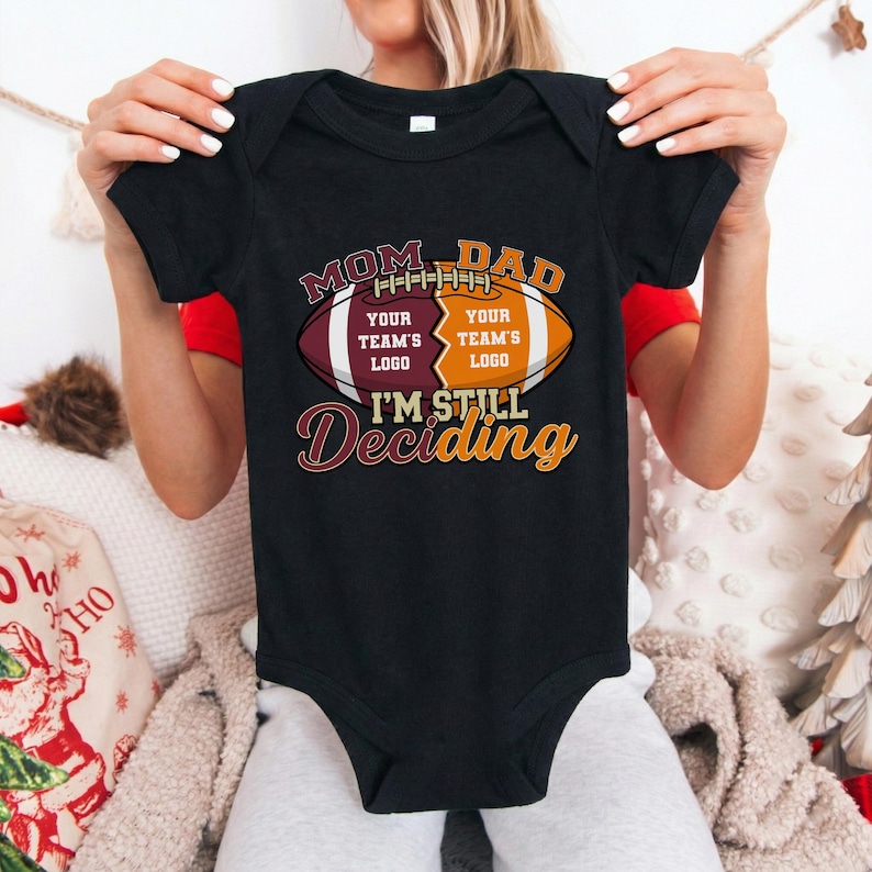 May include: Black baby bodysuit with a football graphic. The graphic reads "MOM DAD" above a football design with space for team logos and the text "I'M STILL Deciding". The bodysuit is held up by a person.