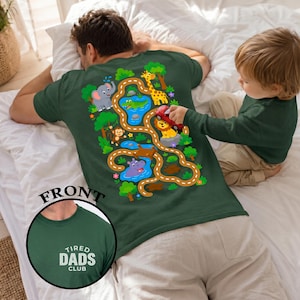 May include: Dark green t-shirt with a colorful jungle-themed design on the back, featuring animals and a road. The shirt has the text "TIRED DADS CLUB" on the front. A child is playing with a toy car on the shirt.