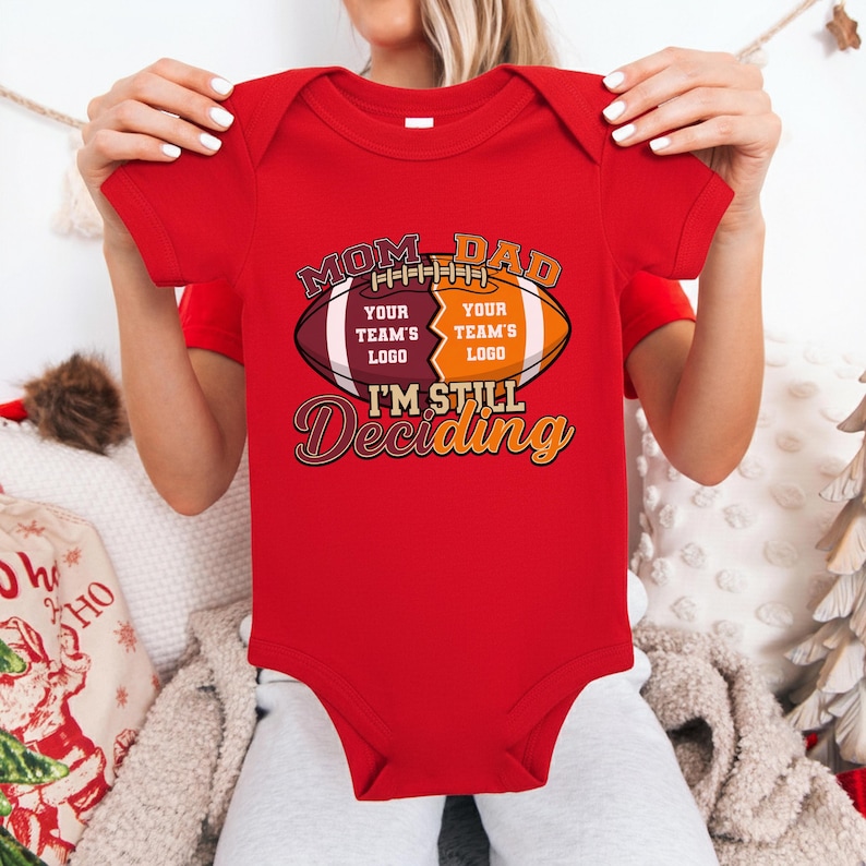 May include: A red baby bodysuit with a football-themed graphic. The graphic includes the words "MOM-DAD", "YOUR TEAM'S LOGO", and "I'M STILL Deciding". The bodysuit is short-sleeved.