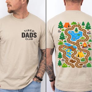 May include: Beige t-shirt with "TIRED DADS CLUB" text on the front. The back features a colorful camping-themed graphic with a road, lake, trees, tents, and a campfire. The design is playful and outdoorsy.