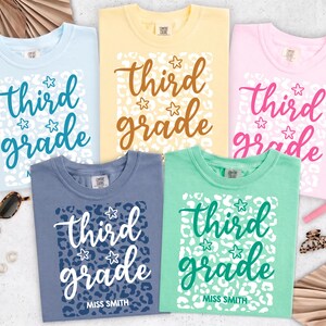 May include: Five crew neck t-shirts in light blue, yellow, pink, slate blue, and mint green. Each shirt features a white leopard print rectangle with the text "third grade" in a cursive font, small star icons, and "MISS SMITH" at the bottom.
