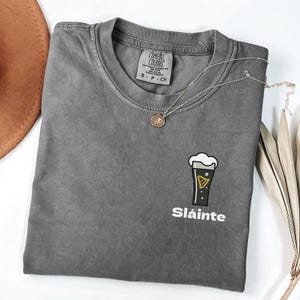 May include: A grey Comfort Colors t-shirt featuring a beer glass design and the word "Sl&aacute;inte". A gold necklace and a brown hat are also visible.
