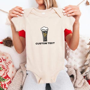 May include: A beige baby bodysuit with a black and yellow graphic of a pint of beer with the text "CUSTOM TEXT" below the graphic. The bodysuit has short sleeves and a round neckline.