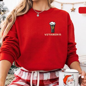 Puede incluir: Una sudadera roja brillante con un gr&aacute;fico de una pinta de cerveza negra y el texto "VITAMIN G". La sudadera de manga larga se usa con pantalones de pijama a cuadros rojos y blancos. Tambi&eacute;n se ve un collar.