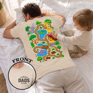 May include: Beige t-shirt with a colorful safari scene on the back, featuring an elephant, giraffe, lion, monkey, and hippo along a road. The front of the shirt has a circular graphic with the text "TIRED DADS CLUB". A child is playing with a toy car.