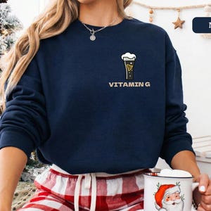 Puede incluir: Sudadera azul marino con un gr&aacute;fico de vaso de cerveza y el texto "VITAMIN G". La sudadera de manga larga y cuello redondo se usa con un pantal&oacute;n de pijama a cuadros rojos y blancos. La persona sostiene una taza.