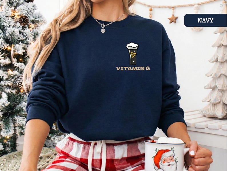 Puede incluir: Sudadera azul marino con un gr&aacute;fico de vaso de cerveza y el texto "VITAMIN G". La sudadera de manga larga y cuello redondo se usa con un pantal&oacute;n de pijama a cuadros rojos y blancos. La persona sostiene una taza.