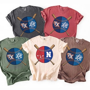 May include: Five short-sleeved t-shirts in various colors, each featuring a baseball design. The baseballs are split in half, with team logos and crossed baseball bats. Colors include gray, red, green, tan, and brown.