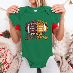 May include: A green baby bodysuit with a football design. The design includes the words "MOM DAD" above a football graphic, with space for team logos. Below the football, the text reads "I'M STILL Deciding".
