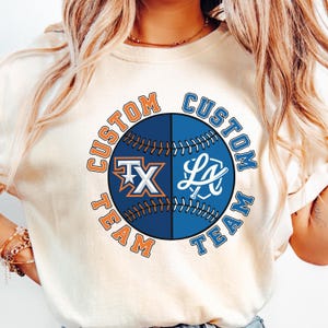 May include: Cream-colored t-shirt featuring a baseball design split in half. One side has an orange and white "TX" logo, the other a white "LA" logo. The words "CUSTOM TEAM" are arched around the baseball in orange and blue.