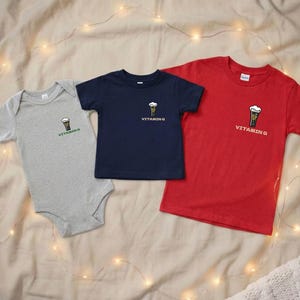 Puede incluir: Tres camisetas y un body para beb&eacute; en gris, azul marino y rojo. Cada prenda presenta un peque&ntilde;o gr&aacute;fico de cerveza y el texto "VITAMIN G". Los art&iacute;culos est&aacute;n colocados sobre una tela de tono neutro.