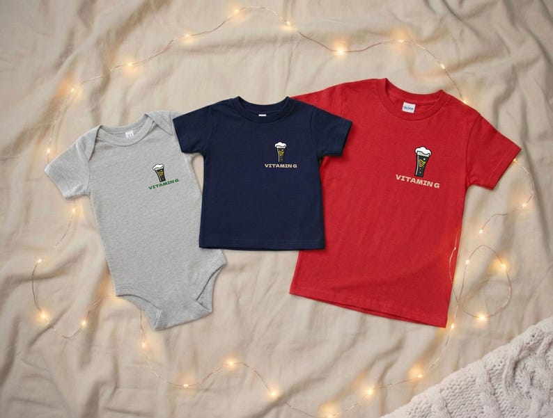 Puede incluir: Tres camisetas y un body para beb&eacute; en gris, azul marino y rojo. Cada prenda presenta un peque&ntilde;o gr&aacute;fico de cerveza y el texto "VITAMIN G". Los art&iacute;culos est&aacute;n colocados sobre una tela de tono neutro.