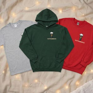 Puede incluir: Tres prendas de vestir: una camiseta gris, una sudadera con capucha verde y una sudadera roja de cuello redondo. Cada art&iacute;culo presenta un peque&ntilde;o gr&aacute;fico de una cerveza con espuma y el texto "VITAMIN G".