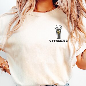 May include: A cream-coloured t-shirt with a graphic of a pint of beer and the text "VITAMIN G". The beer glass is black with a yellow design and white foam. The shirt has rolled-up sleeves.