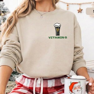 Puede incluir: Una sudadera de color arena con un gr&aacute;fico de una pinta de cerveza y las palabras "VITAMIN G" en verde. La sudadera de manga larga tiene cuello redondo. La modelo lleva un pantal&oacute;n de pijama a cuadros rojos y blancos.