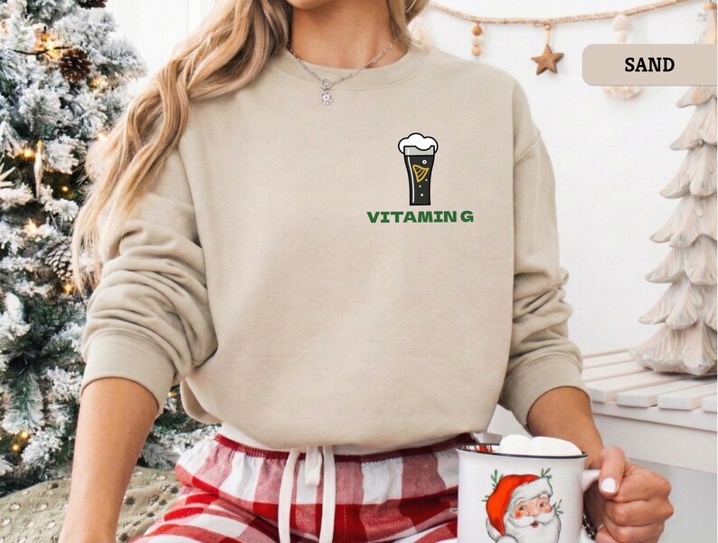 Puede incluir: Una sudadera de color arena con un gr&aacute;fico de una pinta de cerveza y las palabras "VITAMIN G" en verde. La sudadera de manga larga tiene cuello redondo. La modelo lleva un pantal&oacute;n de pijama a cuadros rojos y blancos.