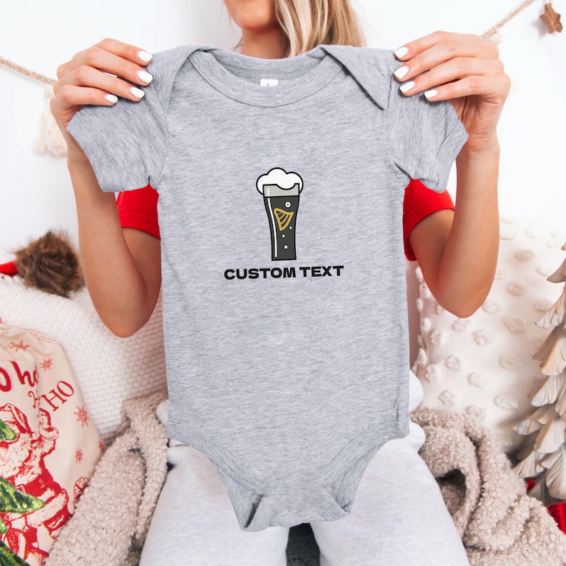 May include: A gray baby bodysuit with a graphic of a pint of stout beer with the text "CUSTOM TEXT" below the graphic. The bodysuit is short-sleeved and has a round neckline.