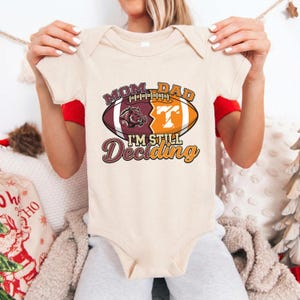 May include: A beige baby bodysuit with a football graphic that reads "Mom Dad I'm Still Deciding." The football design is split between maroon and orange, with a hog and the letter "T" visible.